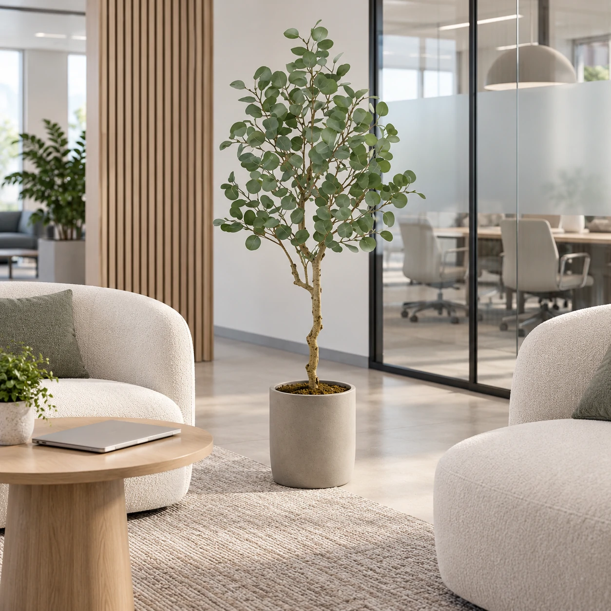 Artificial eucalyptus tree in a gray planter between cream lounge chairs near a glass meeting room.